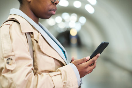 Cropped shot of side view of black woman in beige trenchcoat holding smartphone while texting in...
