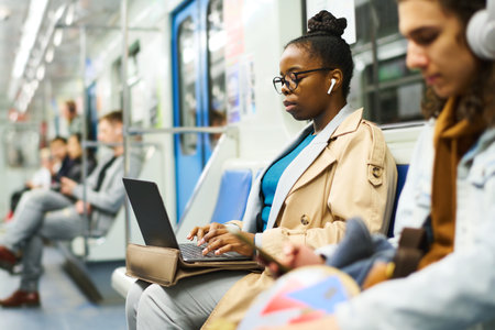 Side view of young serious businesswoman in earphones using laptop while networking or analyzing...