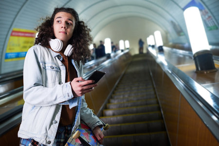 Teenage skateboarder with smartphone standing on moving escalator and looking aside against long tunnel and interior of undergroundの写真素材