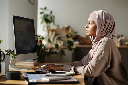 Young designer in hijab looking at computer screen and typing on keyboard while working over new website by workplace at homeの写真素材