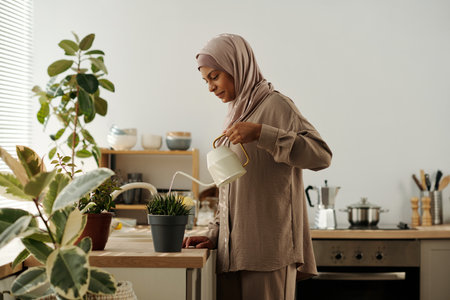 Side view of young smiling Muslim woman in headscarf watering green domestic plants growing in flowerpots standing on kitchen counterの写真素材