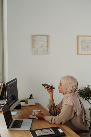 Young businesswoman in hijab recording voicemail on smartphone while sitting by workplace in front of computer screen with coded dataの写真素材