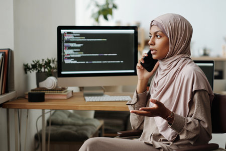 Young confident Muslim female programmer speaking on mobile phone while explaining something to colleague against computer screenの写真素材