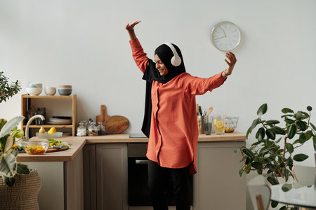 Young cheerful Muslim woman in hijab and headphones dancing in the kitchen in front of counter with fresh chopped vegetables in bowlの写真素材