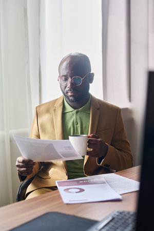 Young serious businessman with cup of coffee looking through financial documents while sitting by workplace in front of camera in officeの写真素材