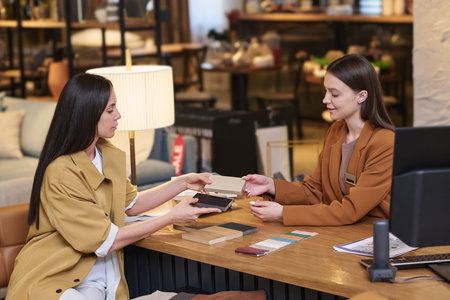 Young smiling designer or manager showing client new facing materials and passing them to her over table with sketches and textile samplesの写真素材