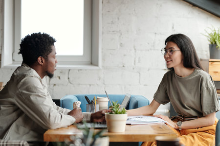 Young smiling businesswoman looking at male colleague during discussion of working points while both sitting by workplace in front of each otherの写真素材