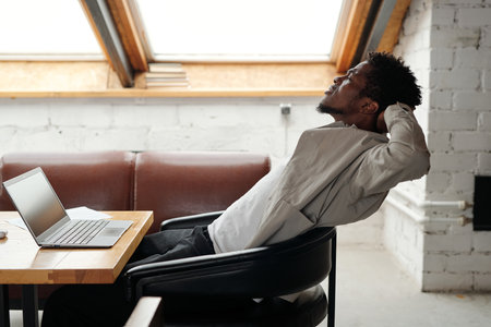Young overworked African American businessman sitting in leather armchair in front of laptop in office and stretching body musclesの写真素材