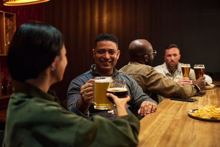 Smiling man with mug of foaming beer toasting with young brunette woman while both standing by...