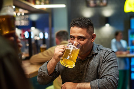 Young man in grey shirt drinking refreshing cool beer from mug while sitting in the bar in front of his buddy and listening to himの写真素材