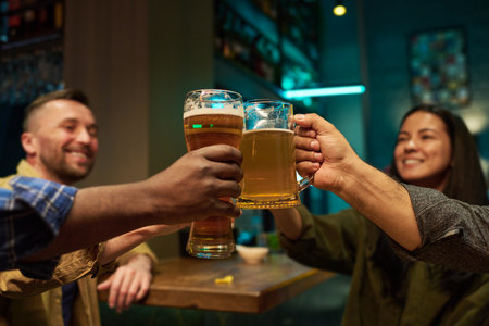 Focus on hands of intercultural buddies clinking with glasses and mugs of refreshing beer while sitting by table and bar counter and toastingの写真素材