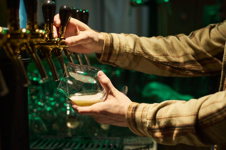 Hands of bartender in checkered shirt pouring fresh lager beer into glass while standing in front of row of faucets and switching one of themの写真素材