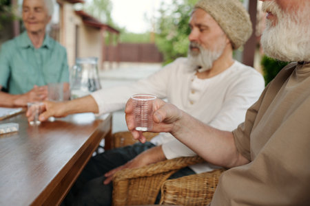 Focus on hand of senior man holding transparent cup with pills while sitting by wooden table in front of camera against other elderly peopleの写真素材
