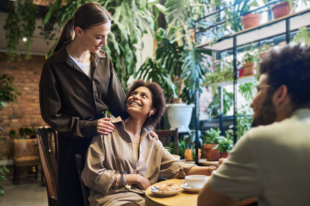 Young brunette woman looking at one of her friends with smile and keeping hands on her shoulders during communication in cafeの写真素材