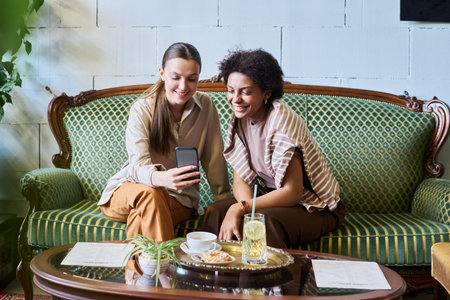 Two intercultural smiling girls looking at smartphone screen while sitting on stylish couch in cozy...