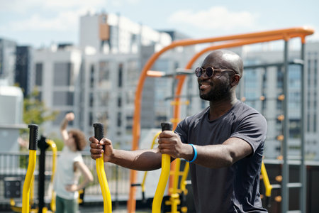 Happy young sportsman in sunglasses and activewear exercising on sports facilities against active woman and urban environment with buildingsの写真素材