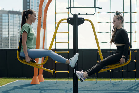 Side view of two young modern sportswomen in activewear sitting on sports facilities in front of...