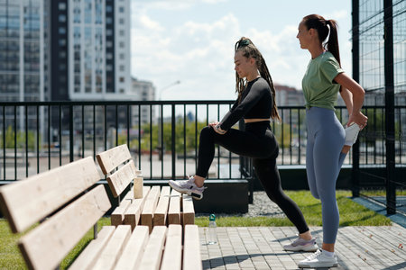 Two young active women in sportswear standing by bench on sports ground on sunny day and doing exercises for leg muscles in open airの写真素材