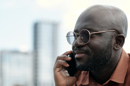 Close-up of young confident African American businessman in eyeglasses holding smartphone by ear while talking to someone outdoorの写真素材