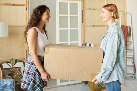 Side view of two happy girls in casualwear carrying big cardboard box and looking at one another during relocation to new apartment or houseの写真素材