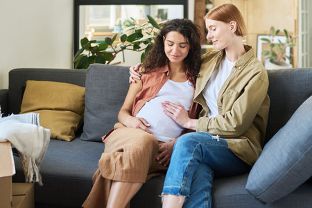 Young smiling woman touching belly of her pregnant girlfriend and embracing her while both sitting...
