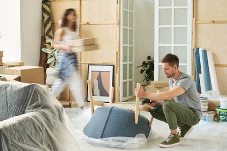 Young man with electric drill repairing wooden details of armchair while sitting on squats against blurry motion of his wife carrying boxesの写真素材