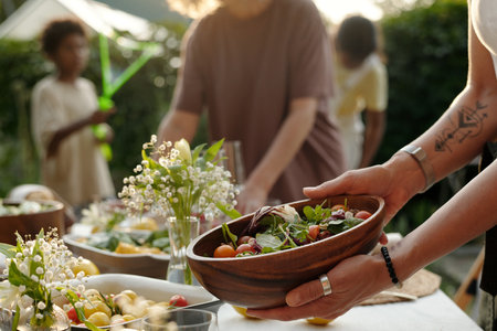 Focus on hands of young woman putting wooden bowl with fresh homemade vegetable salad on table while...