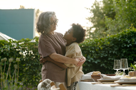 Cute affectionate boy in t-shirt embracing his grandmother and looking at her while both standing against served table before family dinnerの写真素材