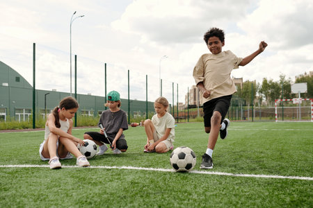 African American schoolboy in activewear running along green football field after soccer ball while playing sports game against his friendsの写真素材