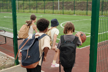 Back view of four schoolchildren entering sports ground or stadium with foorball field while one of them dribbling ball for playing basketballの写真素材