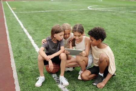 Four youthful intercultural friends in activewear watching online video on screen of tablet held by one of girls while sitting on football fieldの写真素材