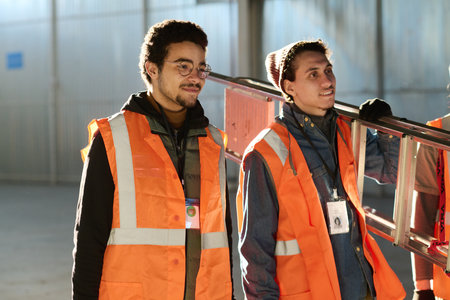 Two young multiethnic warehouse workers in protective fluoresent vests standing in front of camera...