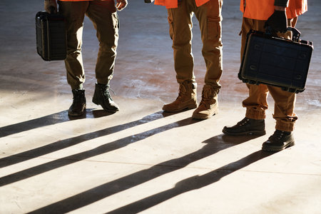 Legs of three male engineers with toolboxes standing on sunlit floor of modern warehouse or industrial plant and having working meetingの写真素材