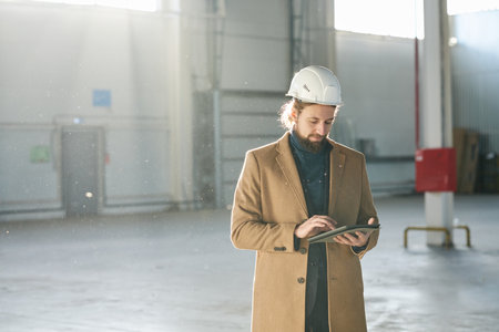 Young bearded builder or solopreneur in hardhat scrolling in tablet while standing in front of camera in spacious sunlit warehouse and networkingの写真素材