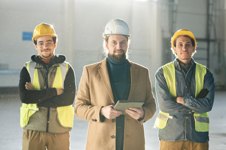 Young successful foreman with tablet standing between two engineers in hardhats and protective fluorescent vests and looking at cameraの写真素材