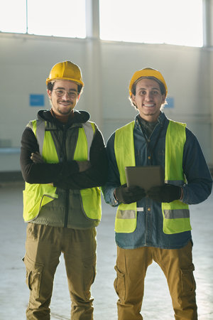 Two young smiling multiethnic engineers standing in front of camera in warehouse lit by sunlight...
