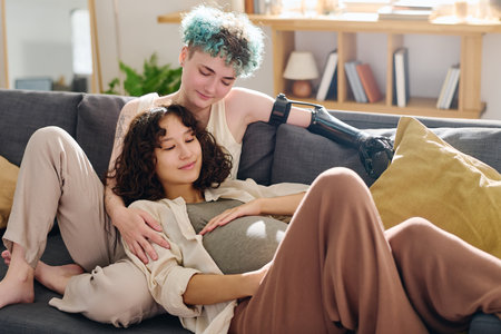 Happy young pregnant woman and her girlfriend with myoelectric hand relaxing on comfortable couch with cushions in home environmentの写真素材