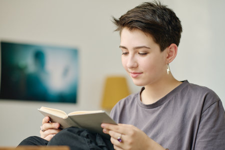 Teenage girl with short dark straight hair looking through text of novel on pages of book in grey...