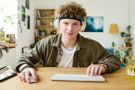 Cute teenage guy keeping hands on mouse and keyboard buttons while sitting by desk in front of computer and looking at data on screenの写真素材