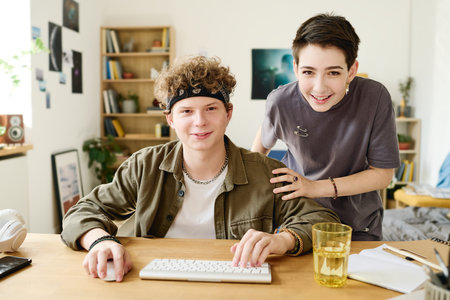 Two happy teenage bloggers in casualwear looking at camera while girl bending over boyfriend typing...