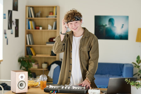 Smiling teenage deejay in headphones and casualwear looking at camera while standing by desk eith turntables and creating new musicの写真素材