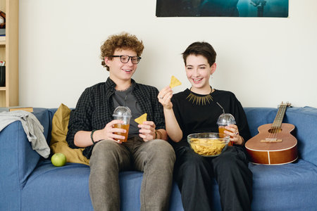 Cheerful teenage couple in casualwear having potato chips with soda while sitting on blue soft couch...