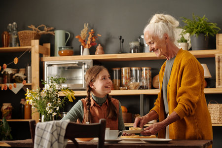 Happy cute girl with pigtails looking at her grandmother while having tea with tasty homemade pie with apples grown in the garden by cottageの写真素材