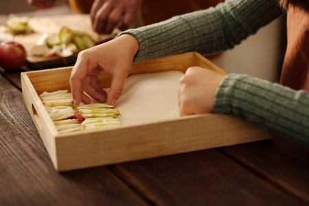 Hands of youthful girl putting slices of fresh ripe apples on wooden tray while helping her...