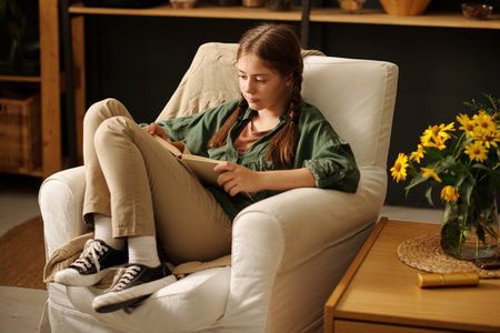 Cute serious schoolgirl in casualwear looking through text on pages of open book while sitting in comfortable armchair in grandmother houseの写真素材