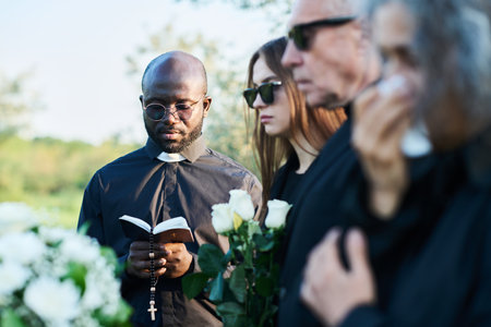 Focus on serious priest in black attire looking in open Holy Bible at funeral service while reading verses aloud and praying in front of coffinの写真素材