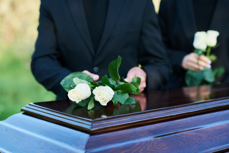 Focus on two fresh white roses being put on coffin lid by mourning mature man wearing black suit standing against young woman with flowersの写真素材