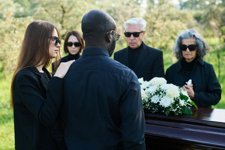 Back view of young woman in mourning attire supporting her husband at funeral while both standing in...