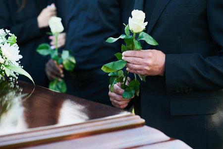 Hand of mature man in mourning clothes holding white rose while standing in front of coffin with...
