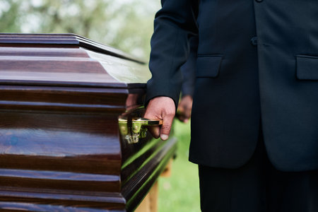 Close-up of man in black suit holding by handle of wooden coffin with dead person while carrying it...